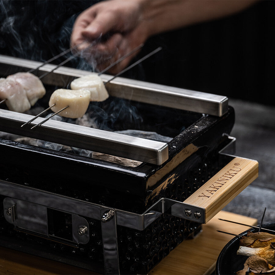 En person griller kjøttspyd over en YAKINIKU Shichirin Yakitori Bars | MEDIUM-grill, med røyk som stiger opp, og fanger den autentiske japanske grillopplevelsen av livlige yakitori-barer.
